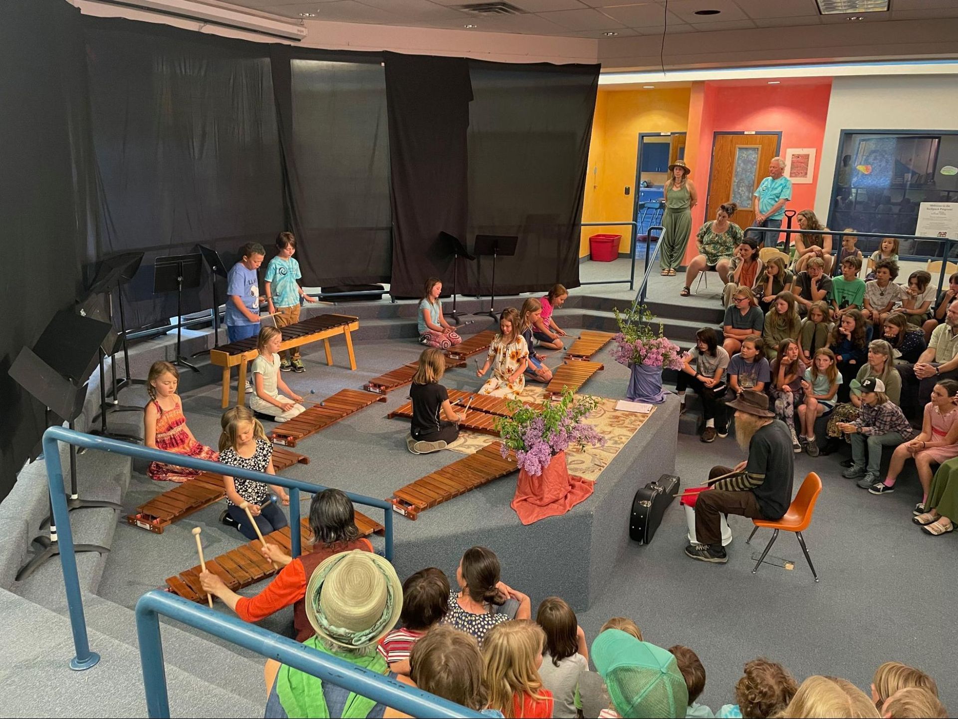 Children performing on a stage in a classroom, with an audience seated around them.