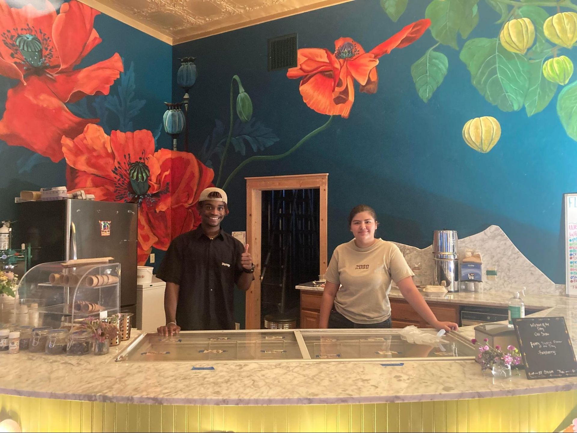 Two people behind a colorful dessert counter with oversized flower wall decor
