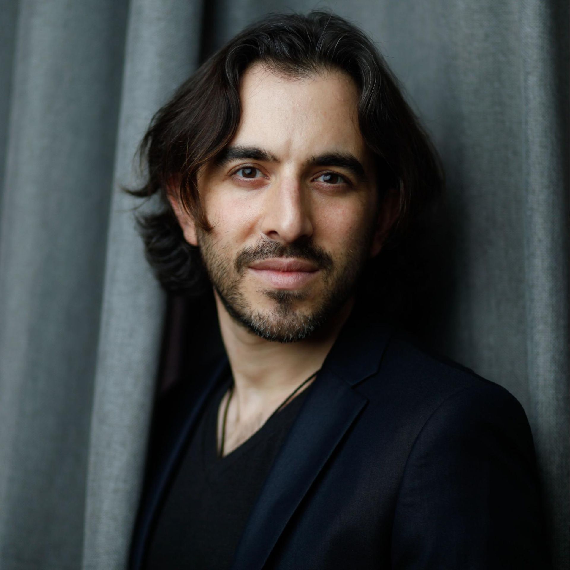 Portrait of a man with dark hair and beard, wearing a black jacket against gray curtains