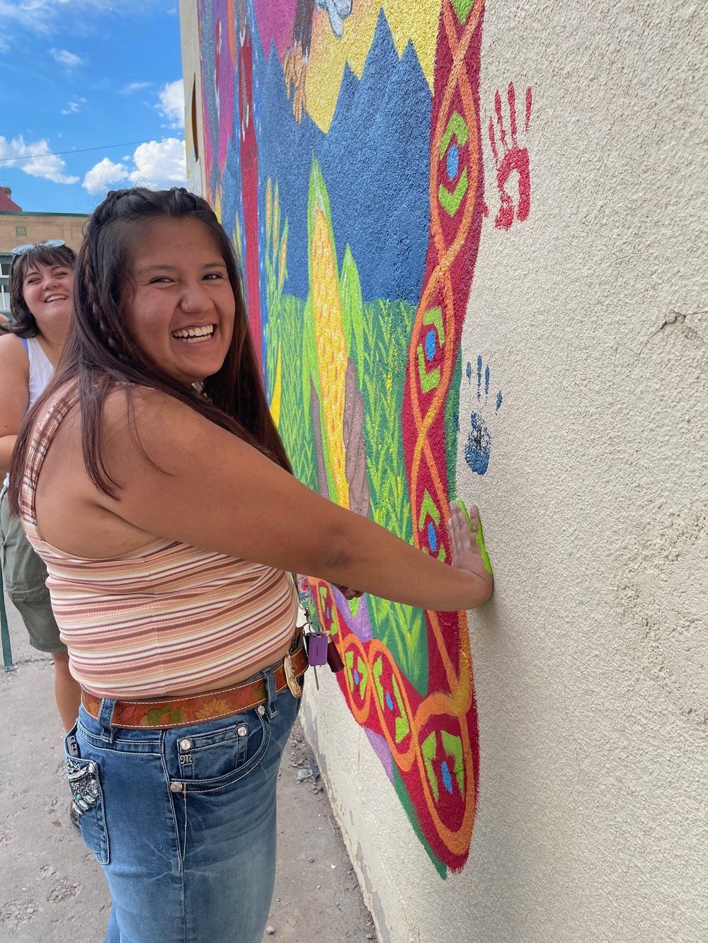 Smiling person painting a colorful mural on an outdoor wall, with another person in the background.
