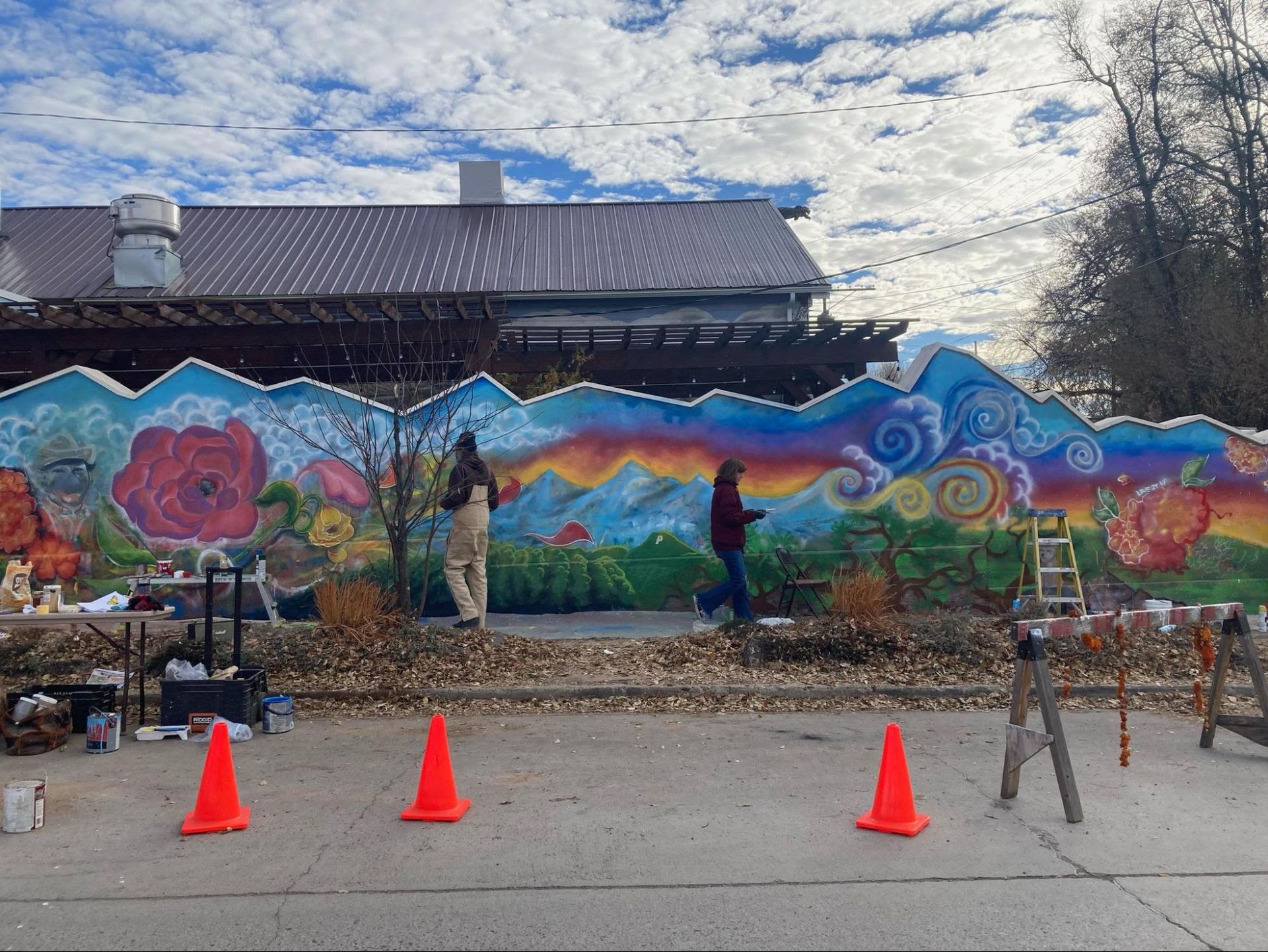 Colorful mural painting outdoors with three orange cones and two people working along a wall