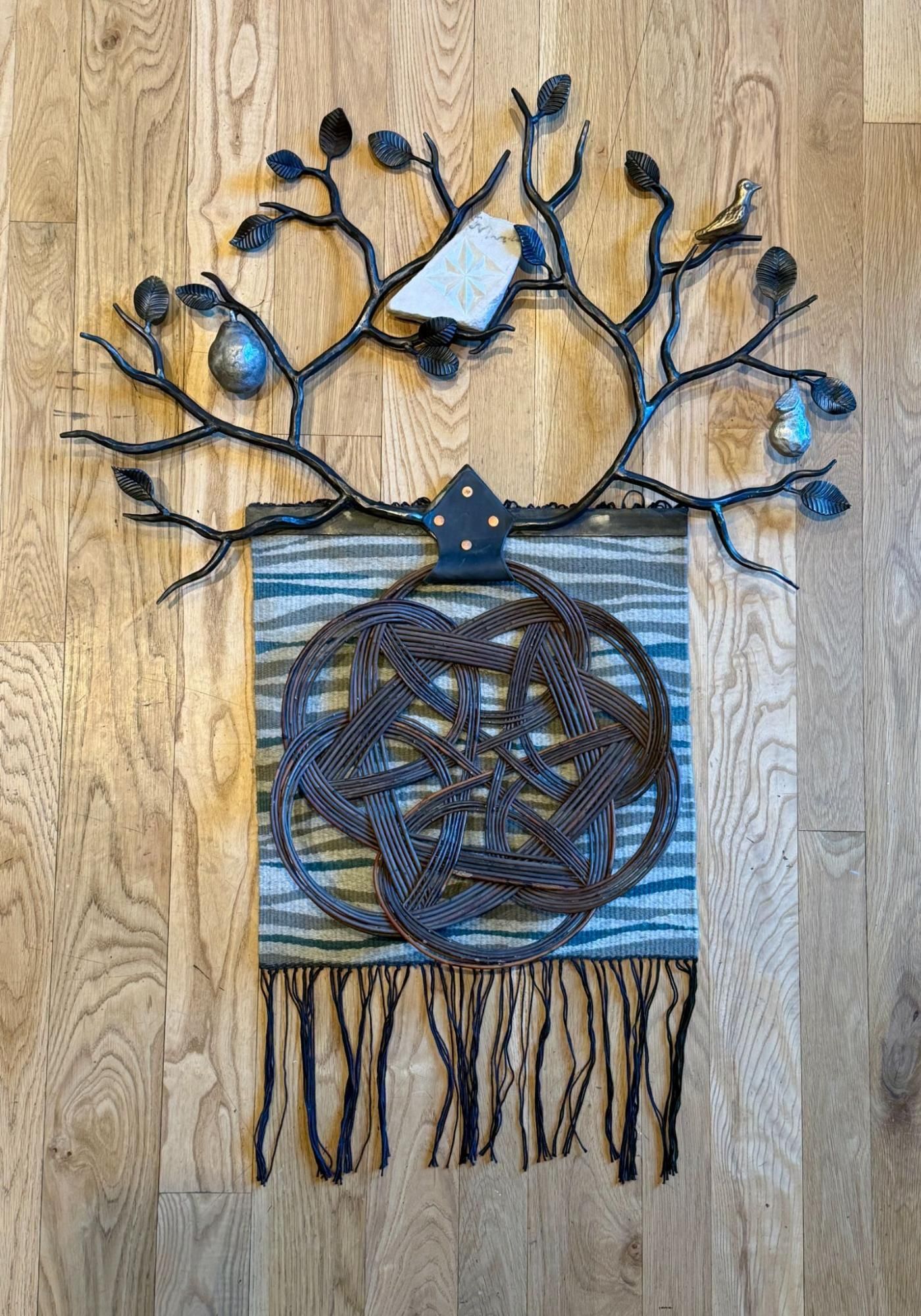 Handmade metal wall art of a tree with birds over a striped woven tapestry and fringe on wood paneling