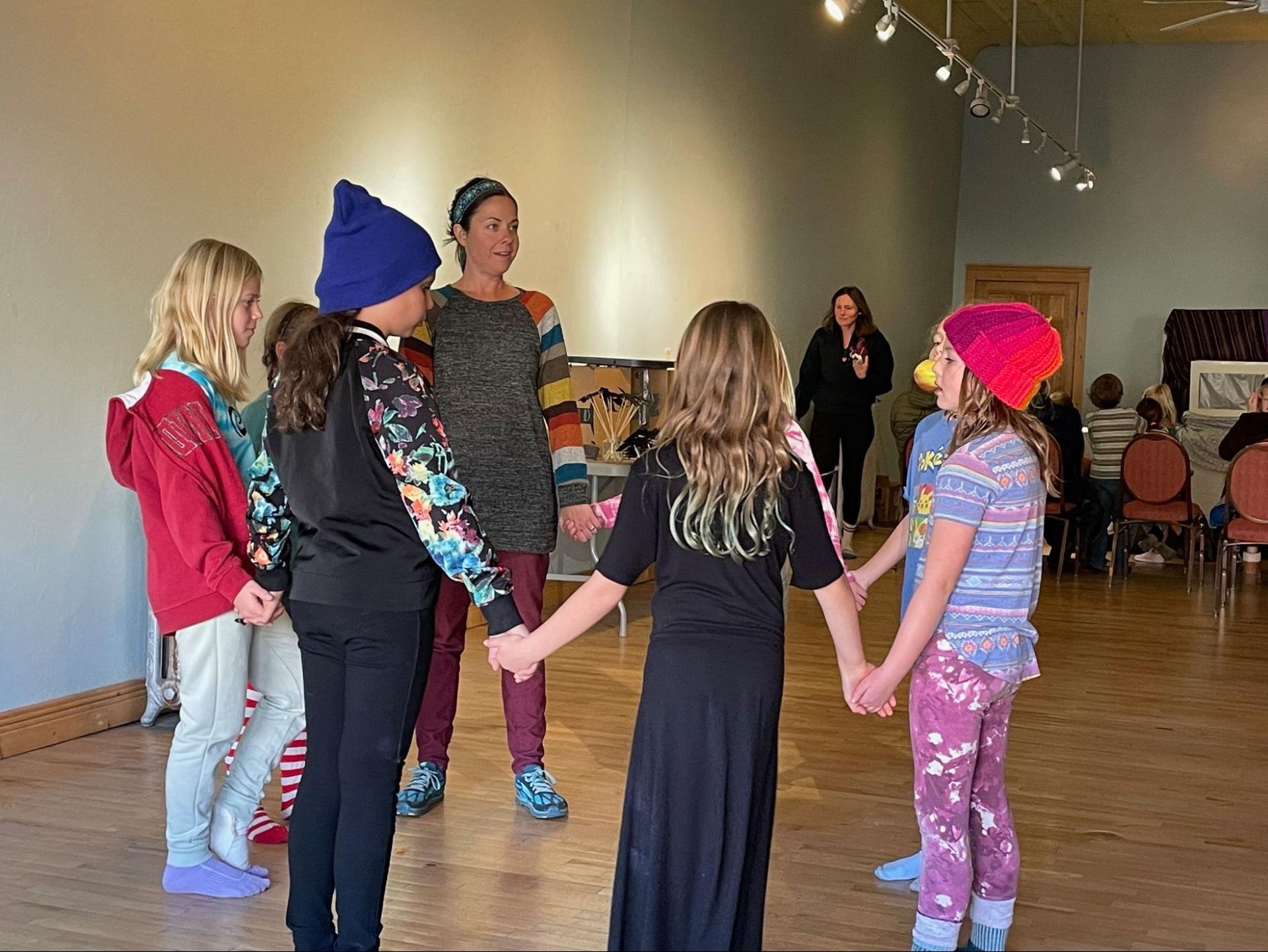 Children holding hands in a circle in a dance studio, wearing colorful hats and casual clothes.
