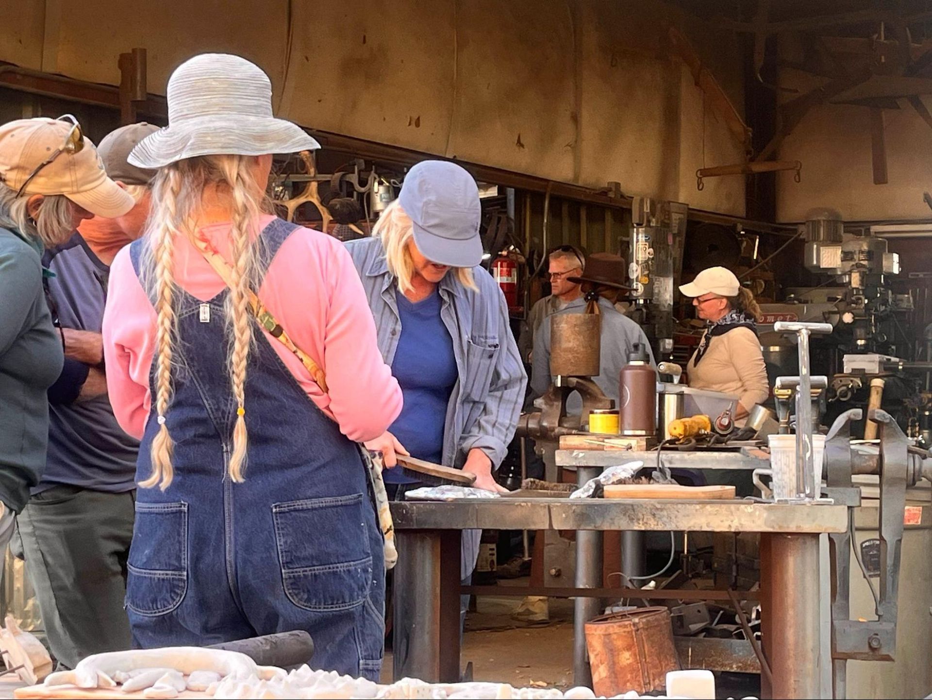 People in hats work at outdoor metal forges in a workshop.