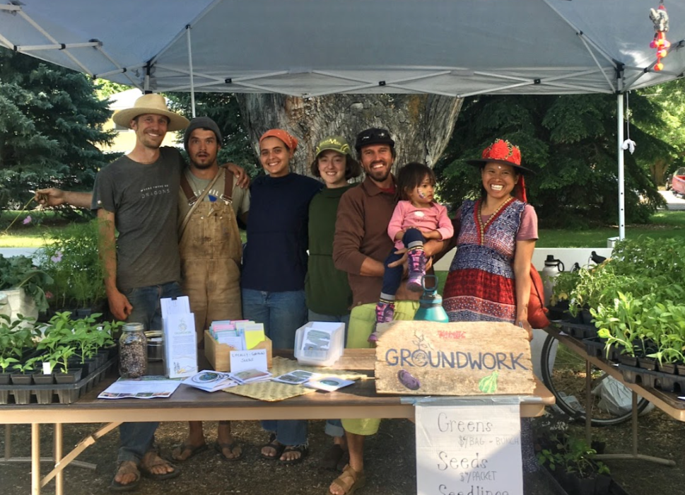 People standing behind a market table under a canopy with plants, signs, and a “Groundworks” banner
