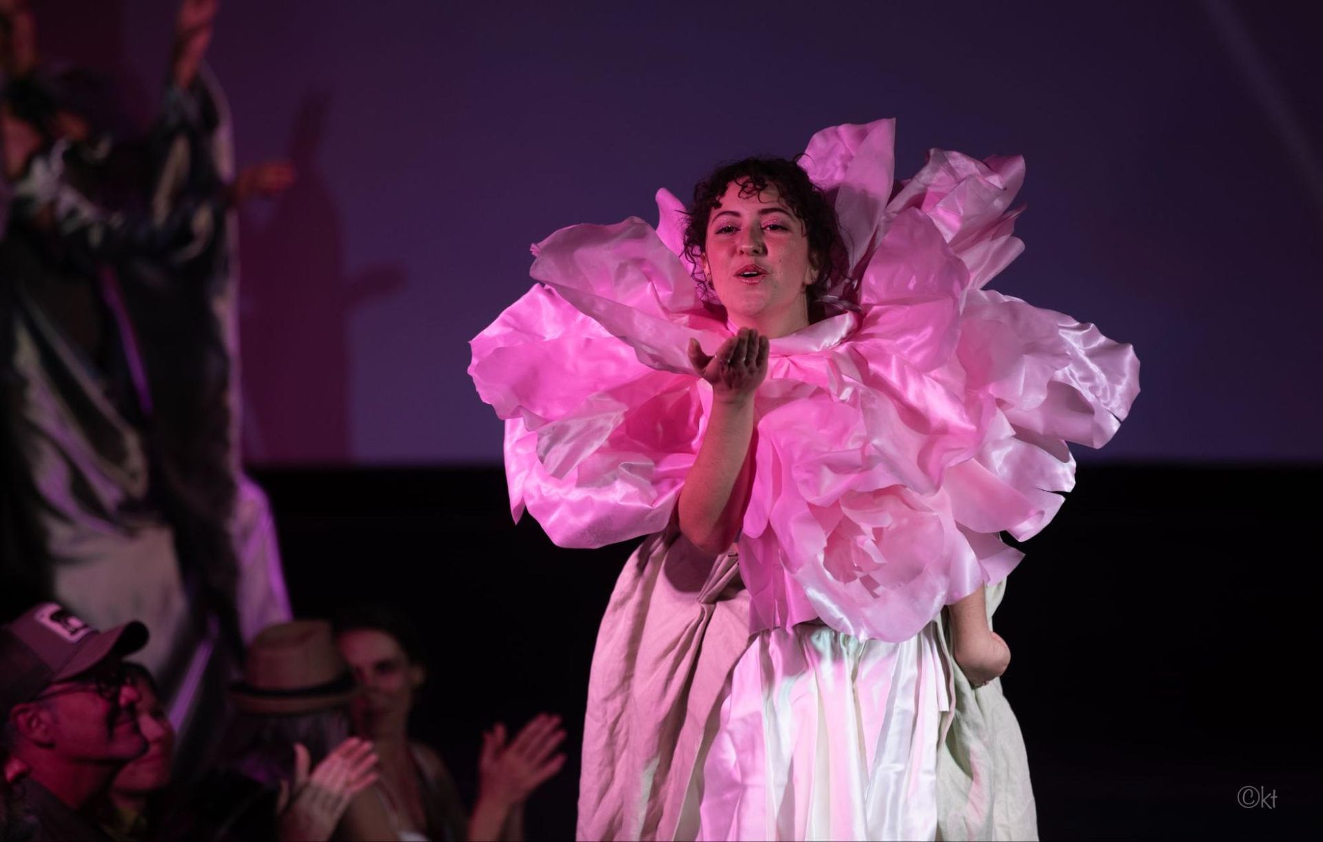 Performer in a pink ruffled dress on stage, blowing a kiss under purple lighting.