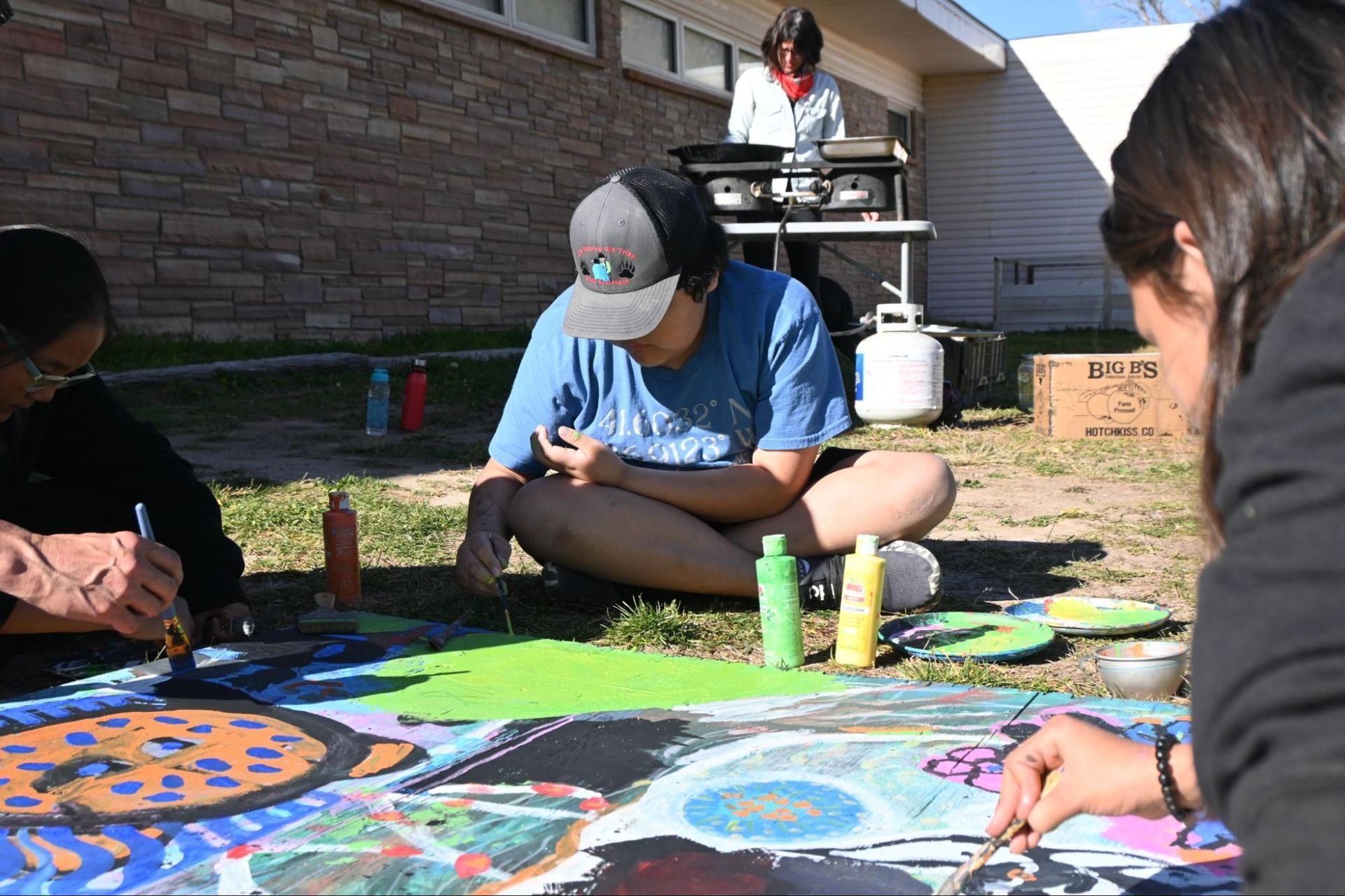 People painting a colorful mural outdoors on large sheets spread across the grass