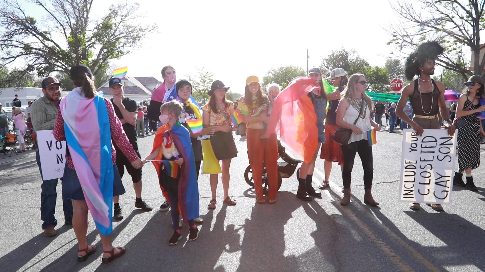 Protesters holding colorful signs and rainbow flags stand across a street in bright daylight.