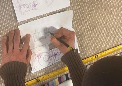 Hands sketching on white paper with colored pencils beside a measuring tape on a woven mat