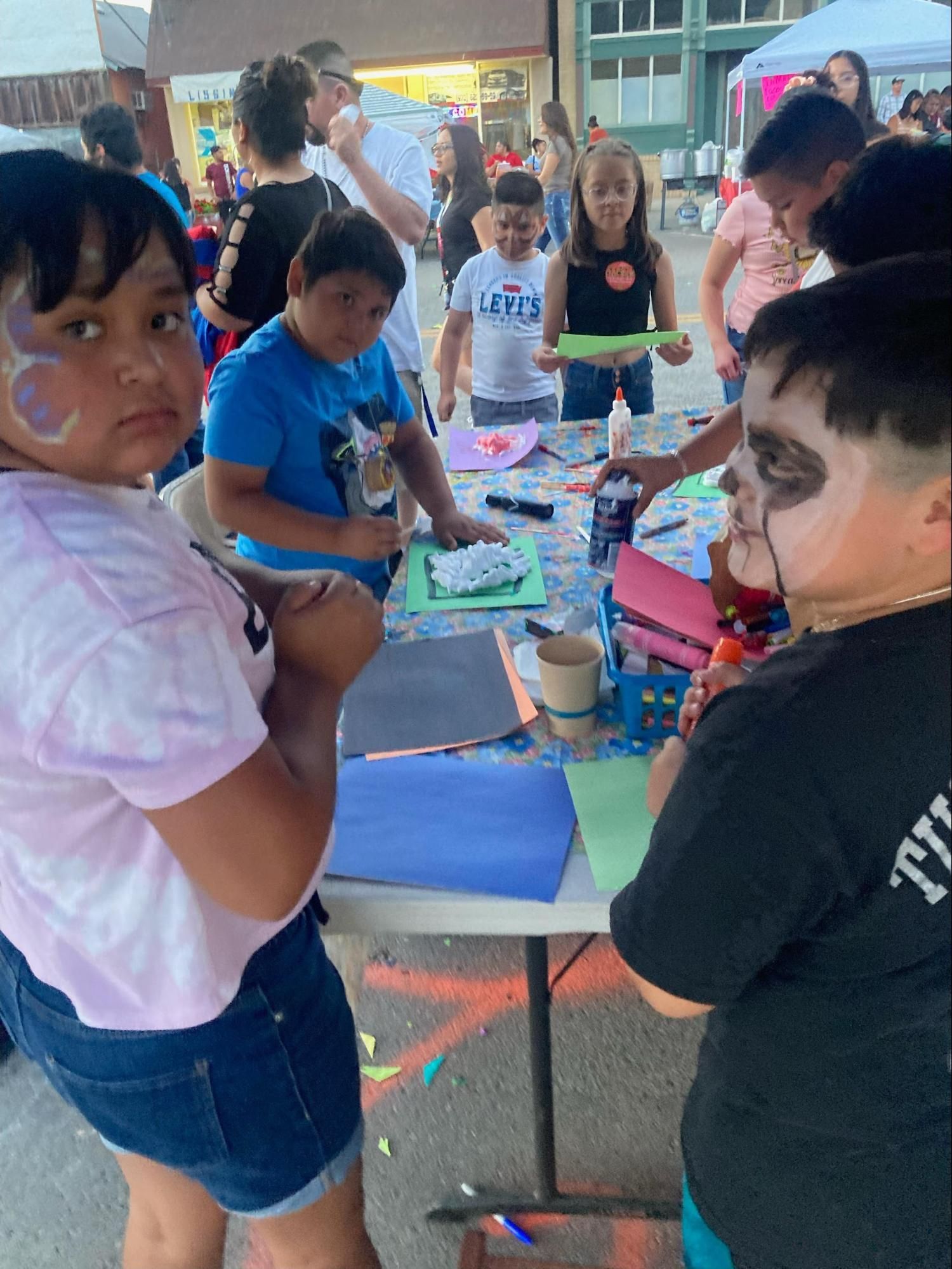 Children gathered around a table doing a craft activity outdoors with markers, paper, and cups.