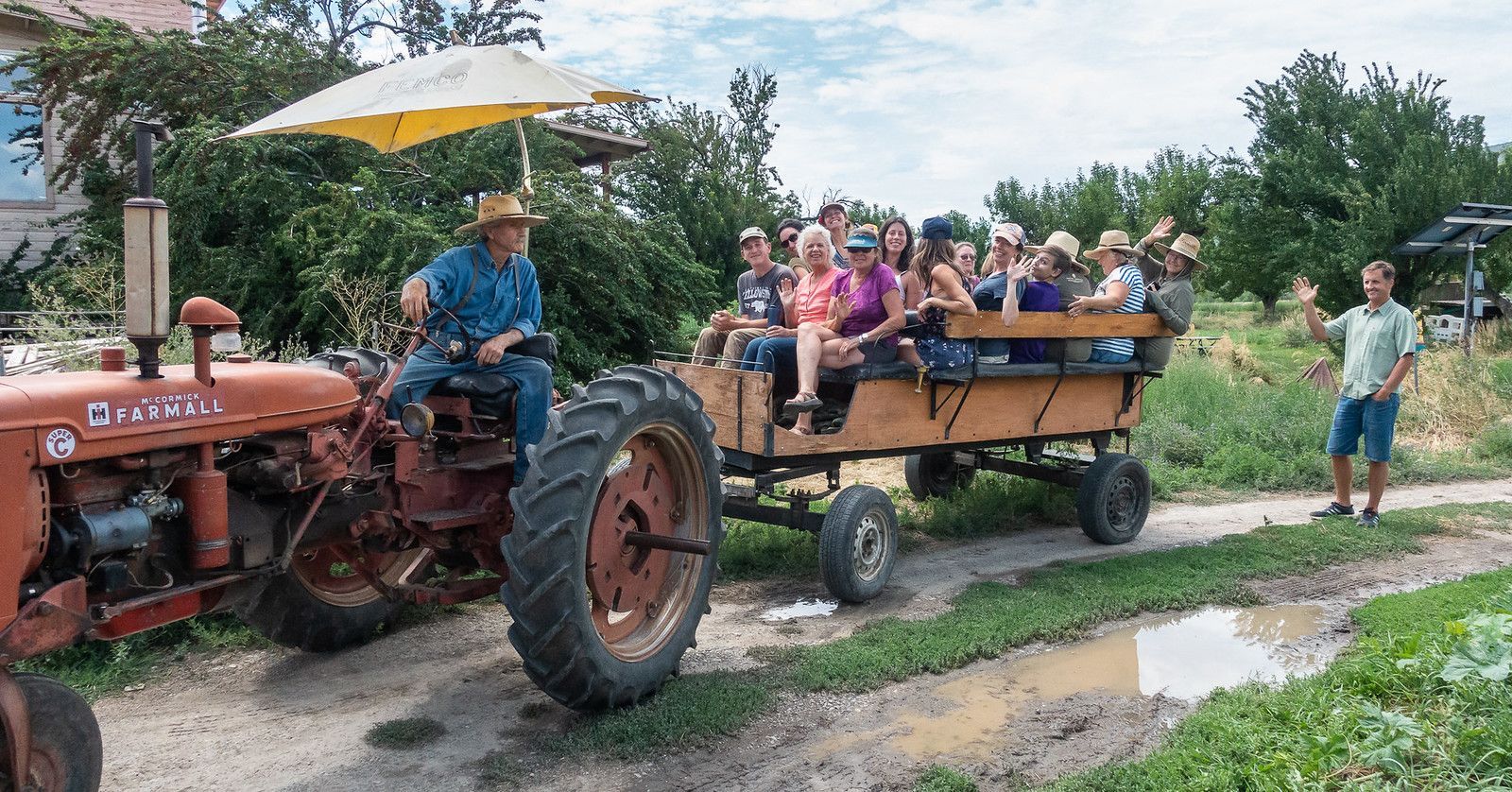 Vintage tractor pulling wagon of seated passengers on a rural dirt road, with a man standing nearby