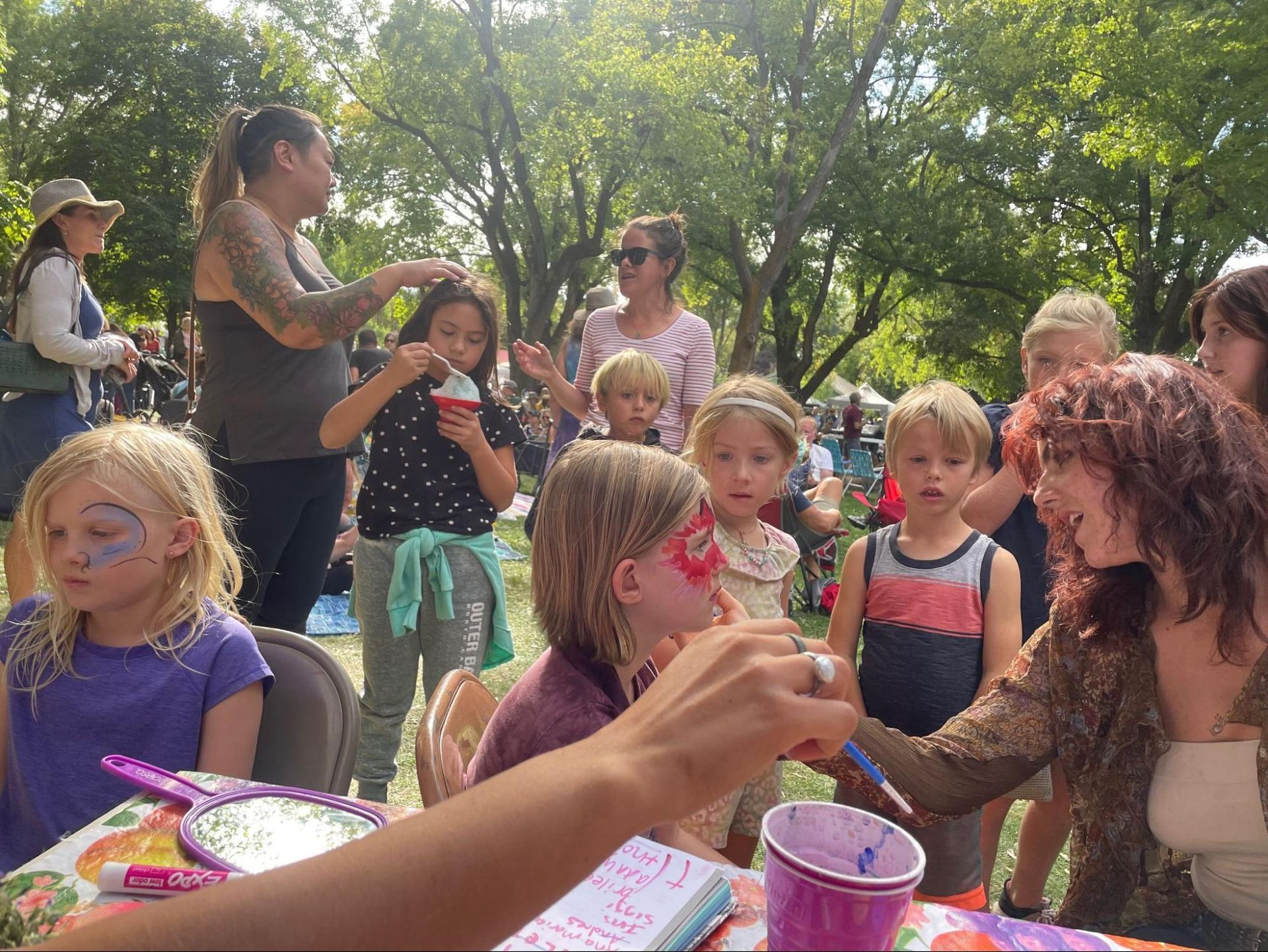 Children gathered outdoors at a party, with cake, snacks, and adults nearby in a shady park.