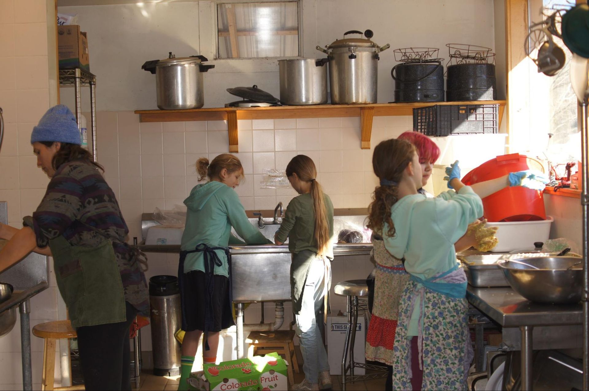 People washing dishes in a busy kitchen with pots, shelves, and a sink.