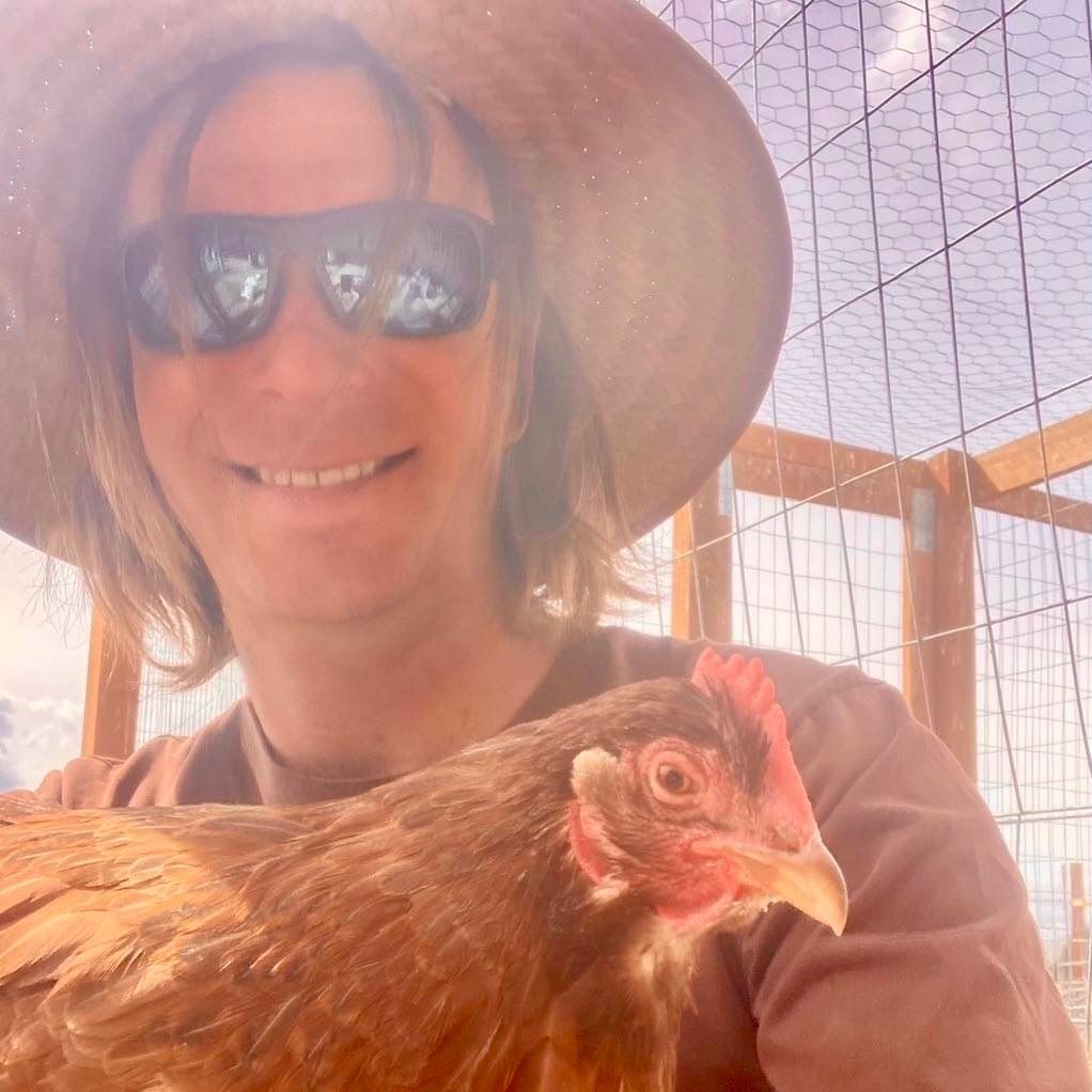 Smiling person in sunglasses and sun hat holding a brown chicken in a fenced outdoor enclosure