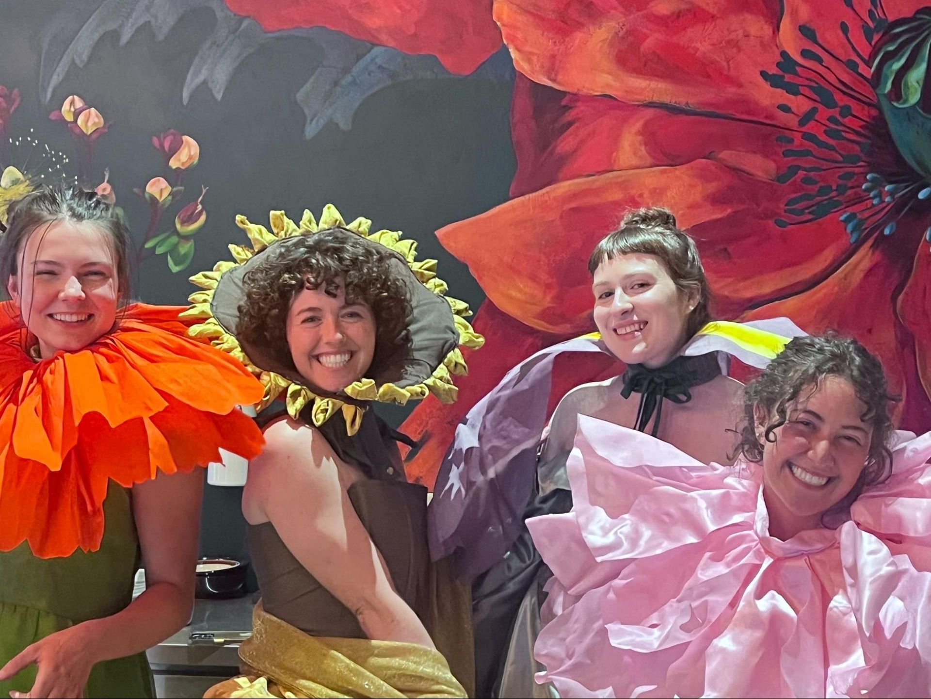 Four people in colorful flower costumes smile in front of large painted flowers.