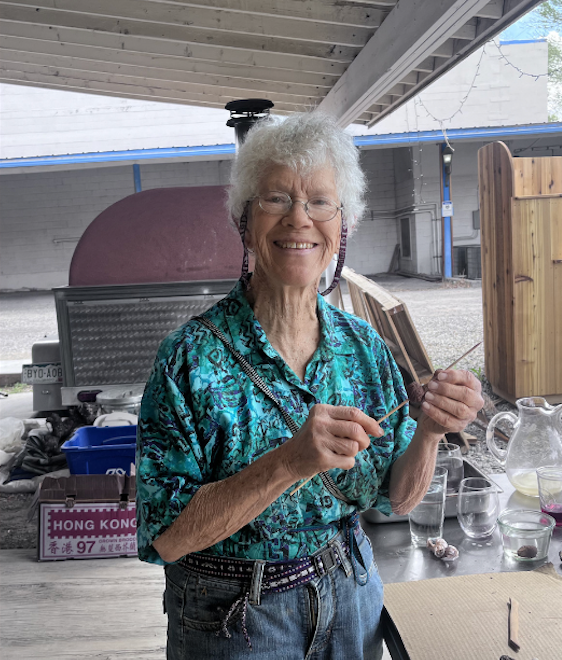 Smiling person in teal blouse holding utensils in a covered outdoor kitchen area