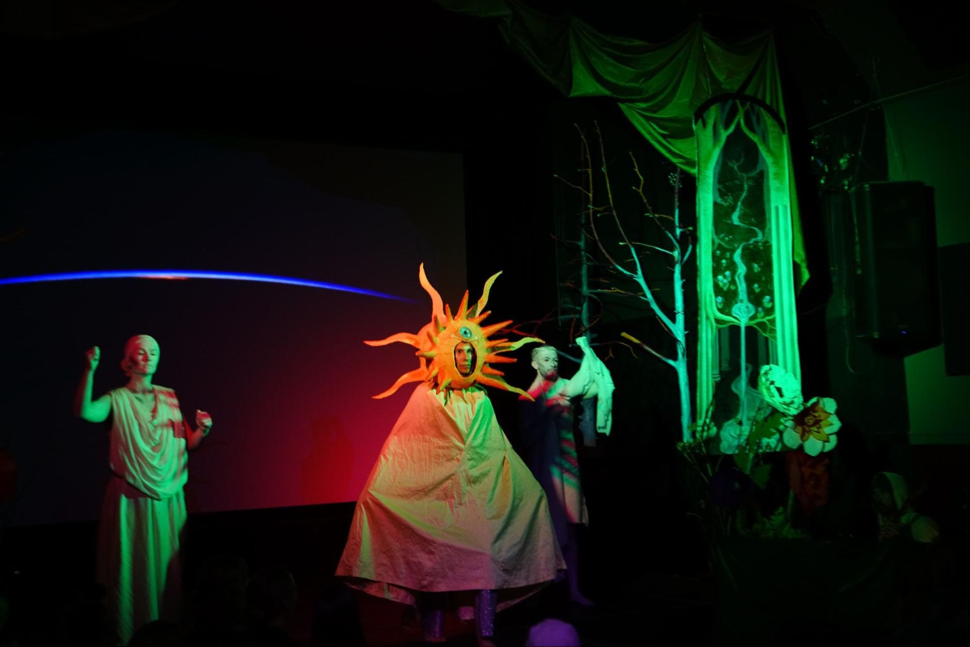 Theatrical stage scene with performers in white costumes under green and red lighting, centered on a glowing sun mask