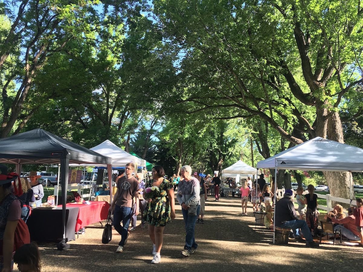 Shaded outdoor market with white tents and people browsing along a tree-lined path