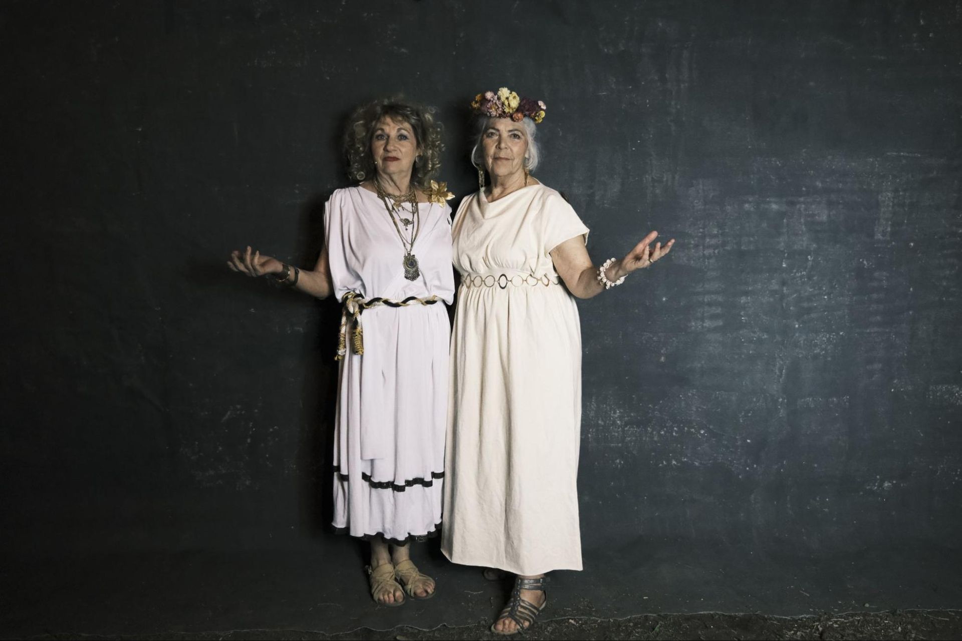 Two people in white dresses standing before a dark wall, arms raised in a welcoming pose.