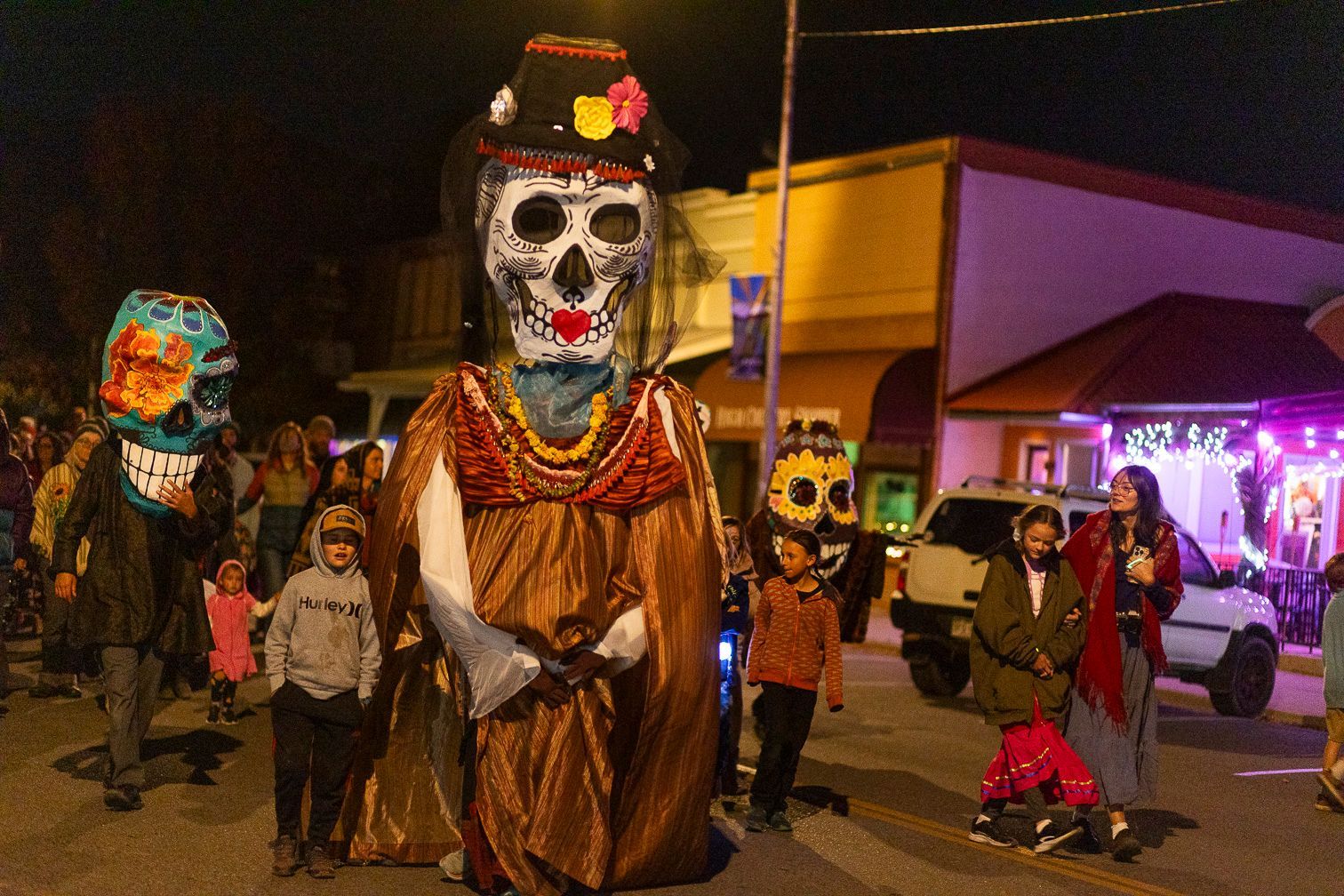 Night parade with giant skull figures and people walking through a brightly lit street at dusk