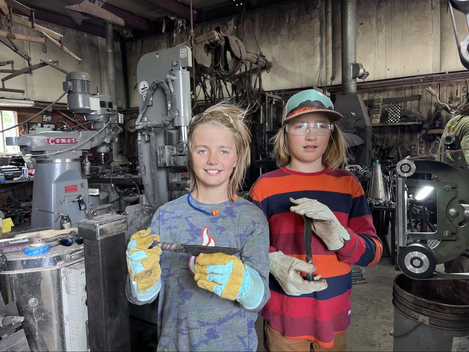 Two workers in gloves and safety glasses standing in a workshop with industrial machinery and tools.