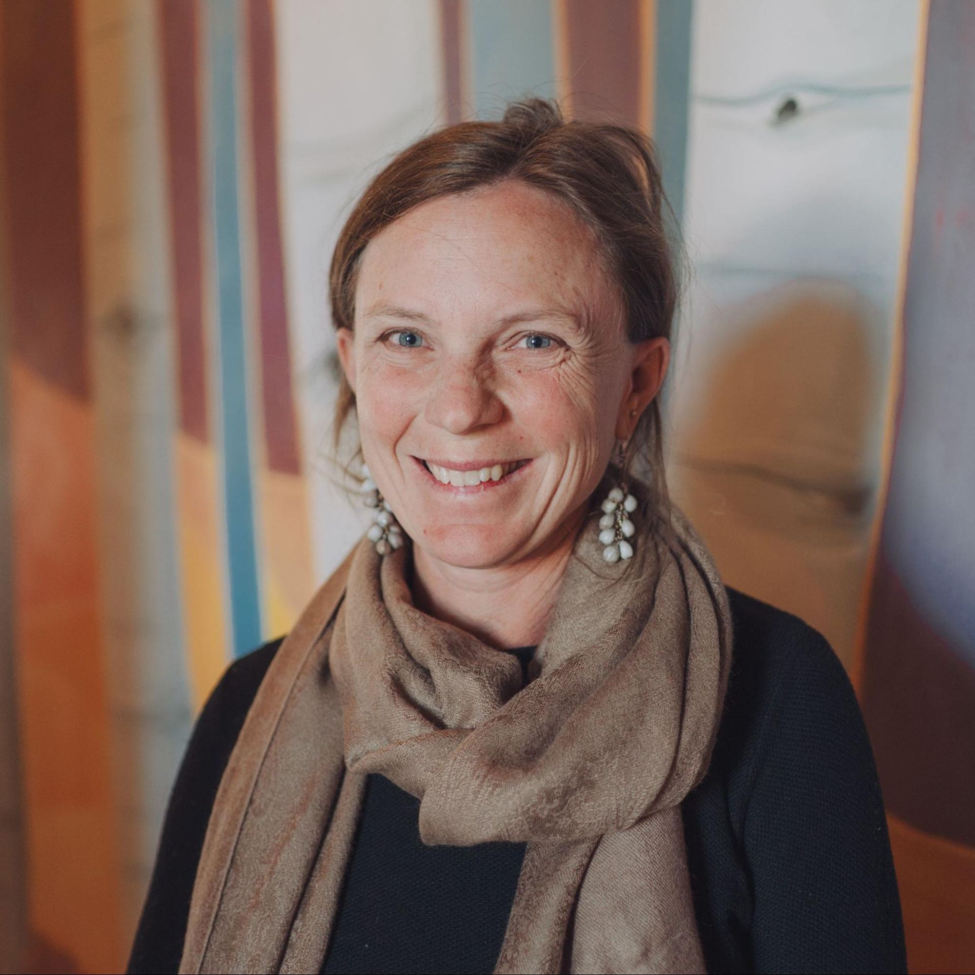 Smiling woman in a beige scarf and earrings, standing indoors against a softly blurred striped background