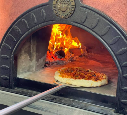 Pizza baking in a wood-fired oven, with flames burning in the back and a pizza peel holding it in front