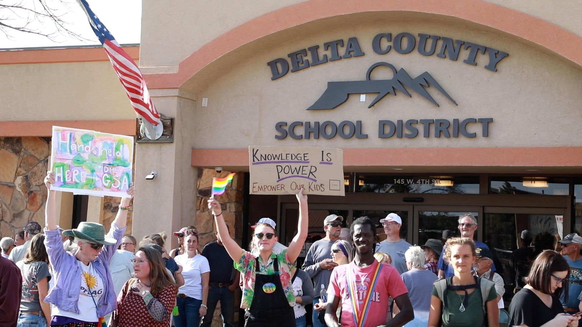 Protesters holding signs outside Delta County School District building with an American flag overhead