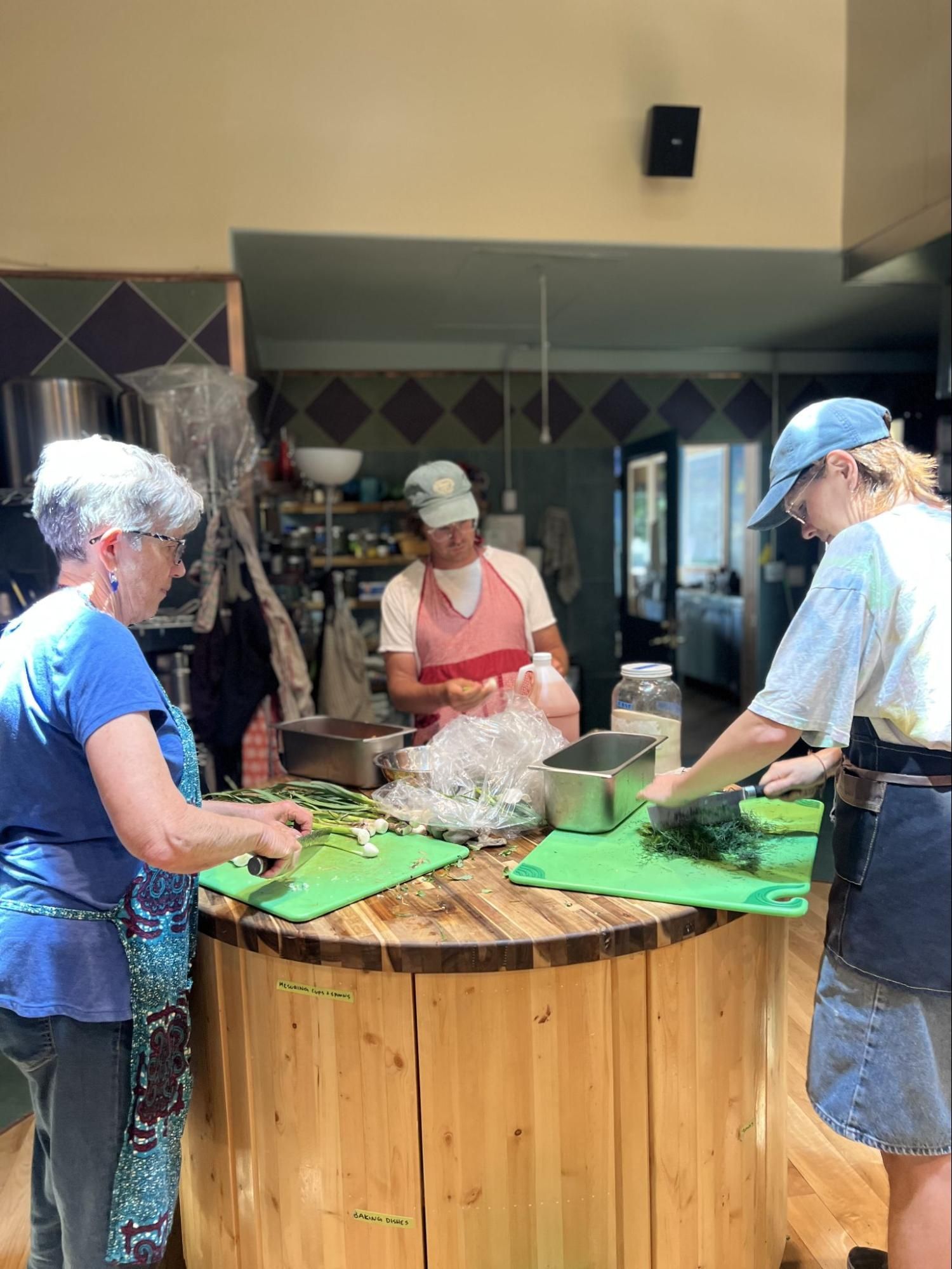 Three people preparing food at a round wooden counter in a kitchen, chopping ingredients on green cutting boards.
