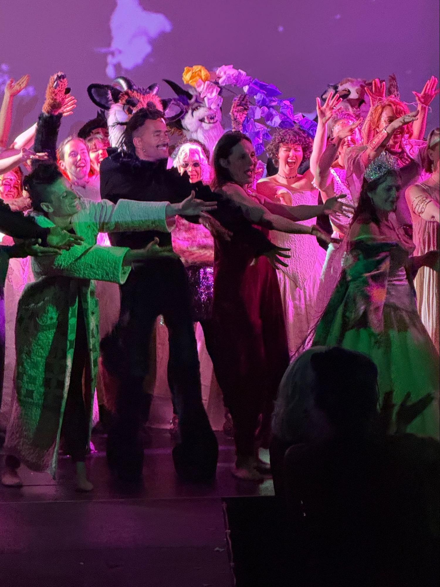 Dancers in colorful costumes perform onstage under pink and purple lighting.