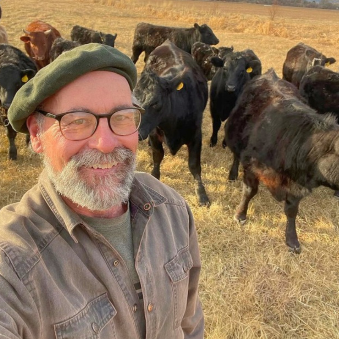 Smiling man in a green beret stands in a dry field with black goats behind him.
