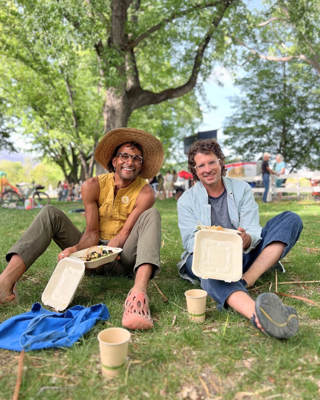 Two people sitting on grass under a tree, smiling with buckets and a blue blanket in a park.