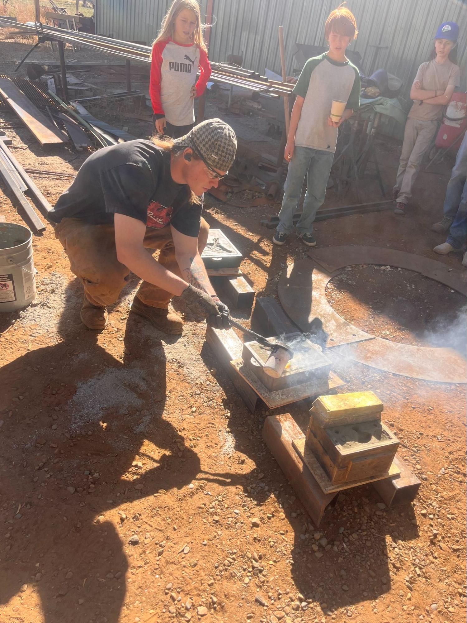 Person in black shirt using a shovel beside a smoking fire pit outdoors, with bystanders nearby.