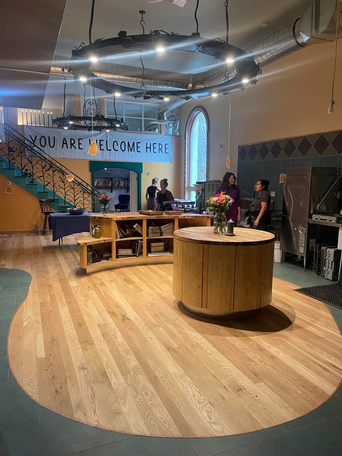 Indoor lounge with wooden floor, round table, booths, and people near a counter under a “YOU ARE WELCOME HERE” sign
