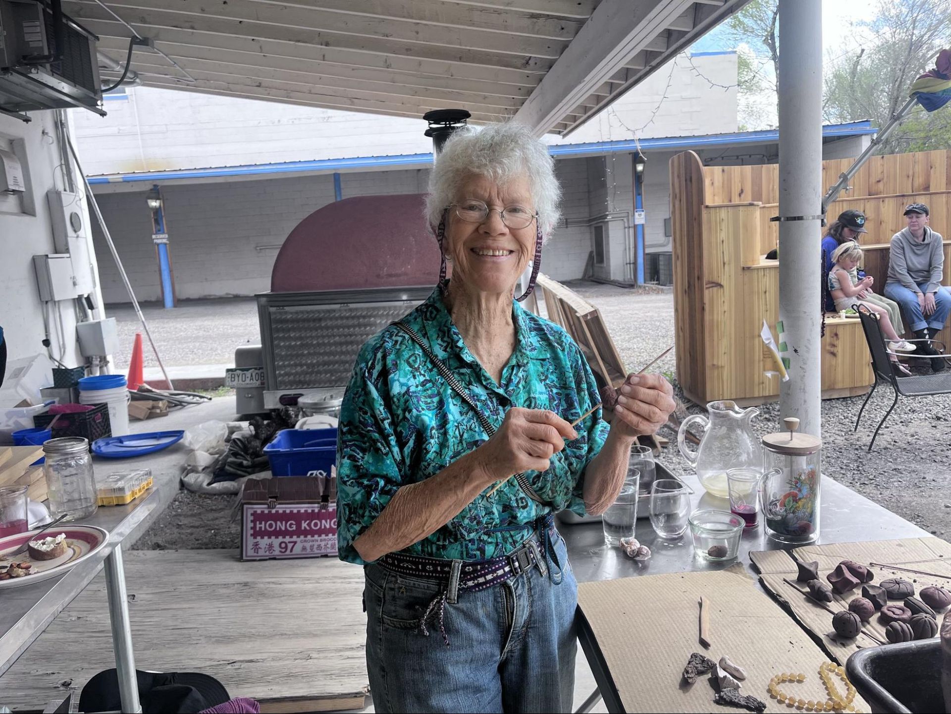 Person in a teal shirt holding skewers beside a barbecue grill on a covered patio.
