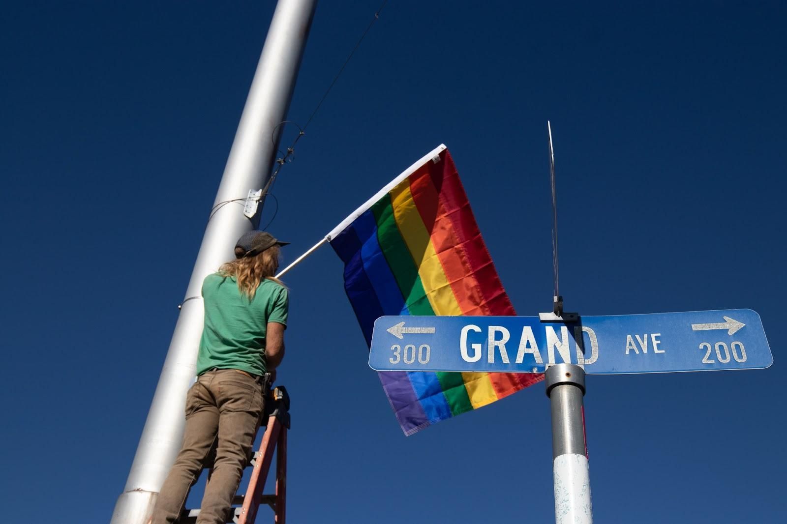 Person on ladder attaching a rainbow Pride flag to a street sign under a clear blue sky
