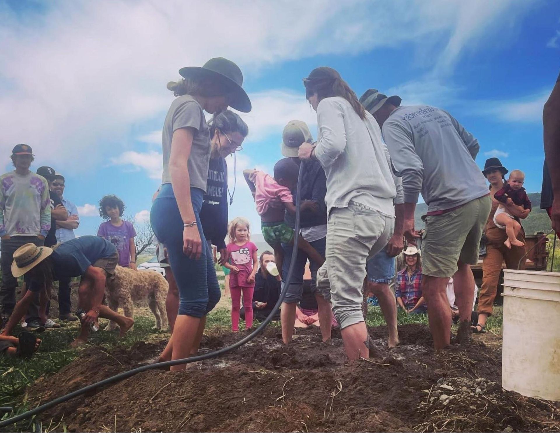 People gathered outdoors around a muddy garden plot, working with shovels under a blue sky.