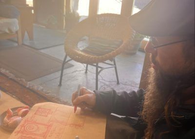 Person writing in a notebook at a table in a sunlit café, wearing a hat and glasses.