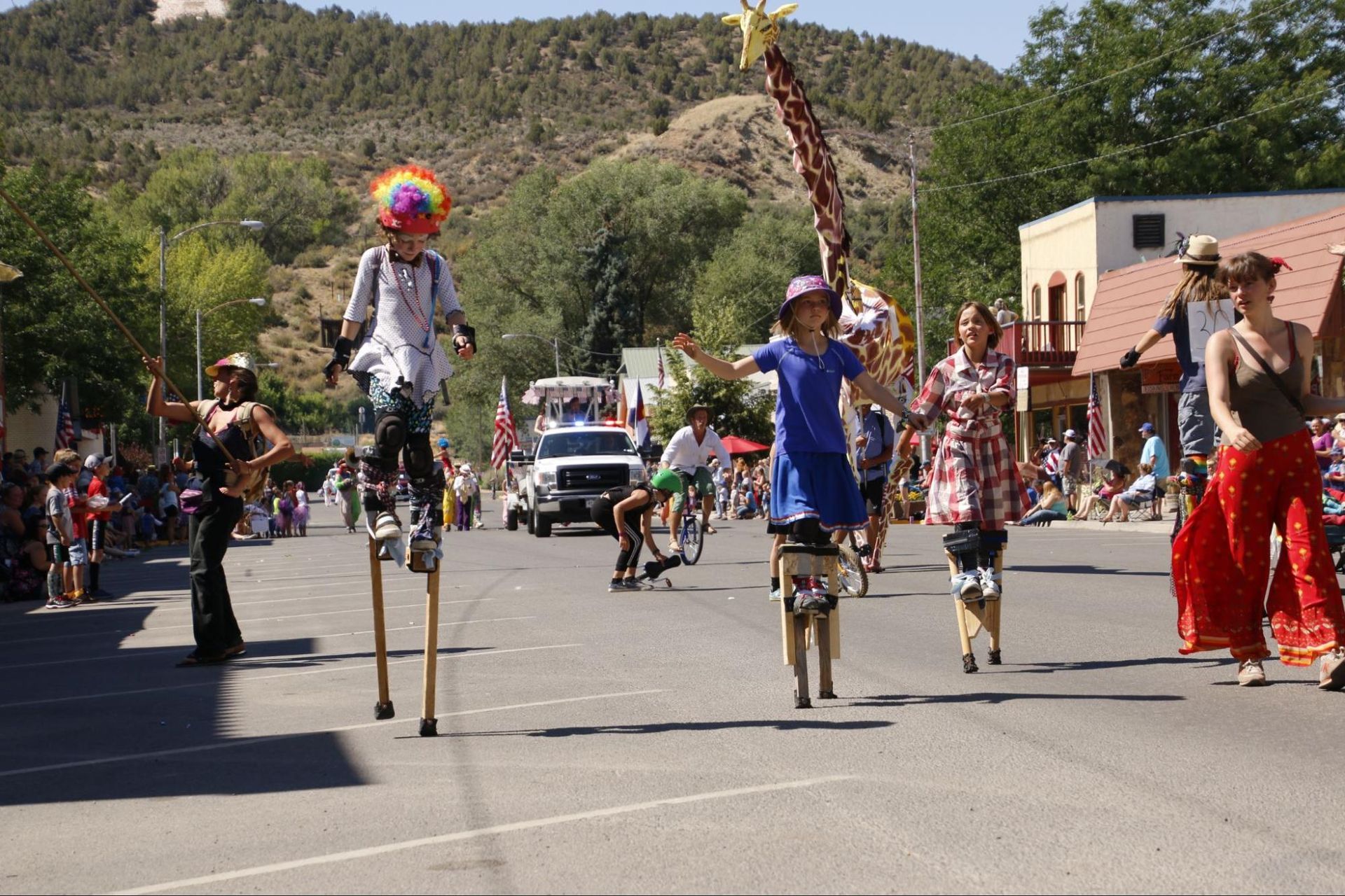 Street parade with stilt walkers in colorful costumes marching down a road, with spectators and mountains behind.