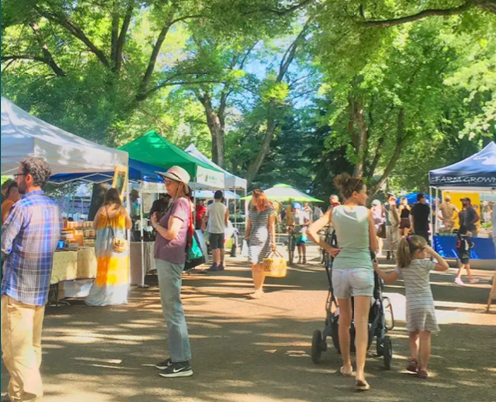 People browsing outdoor market stalls under trees on a sunny day