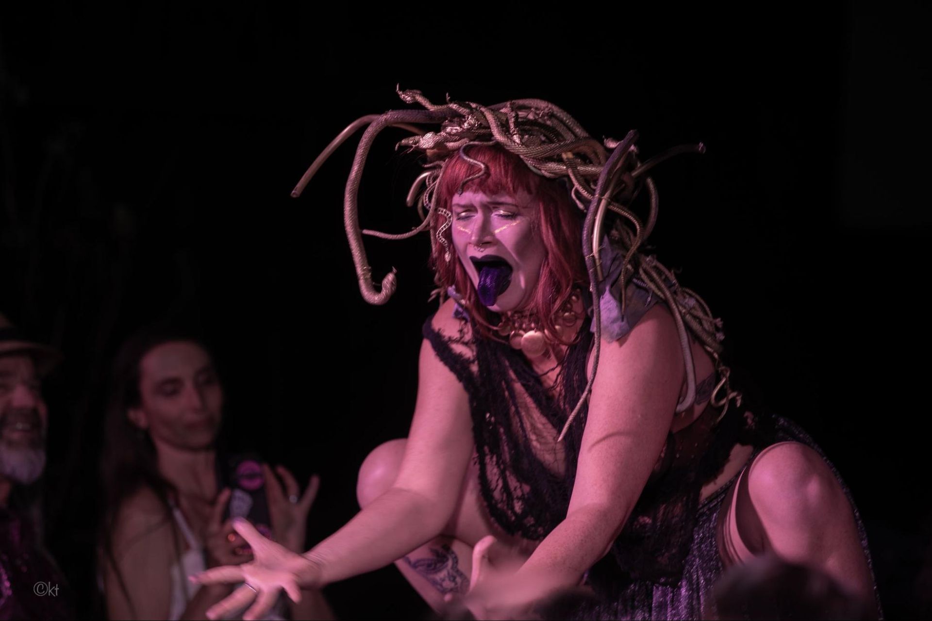 Performer in a pink wig and dark costume, crouching and shouting on a dim stage under pink lighting