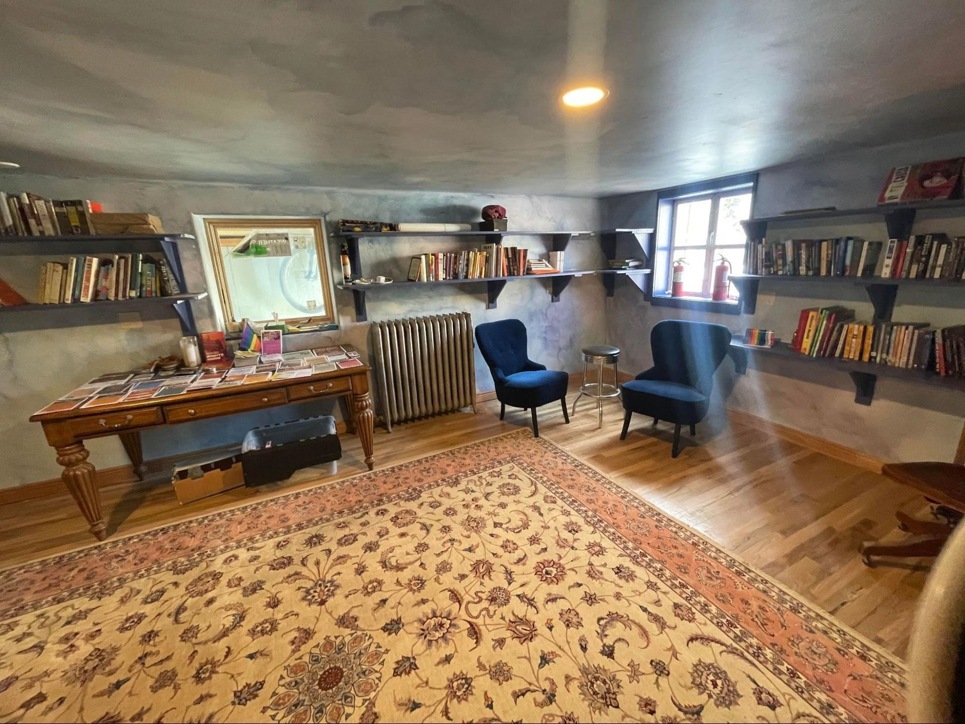 Cozy living room with patterned rug, blue chairs, bookshelves, desk, and a window seat