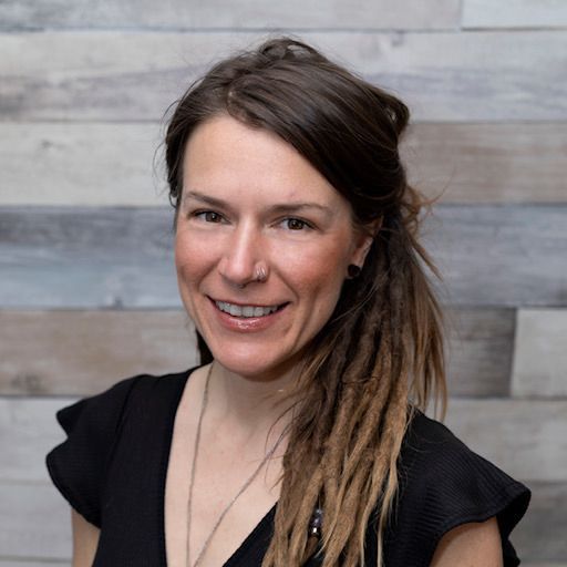 Smiling person with brown hair in a black top against a gray wood-paneled background