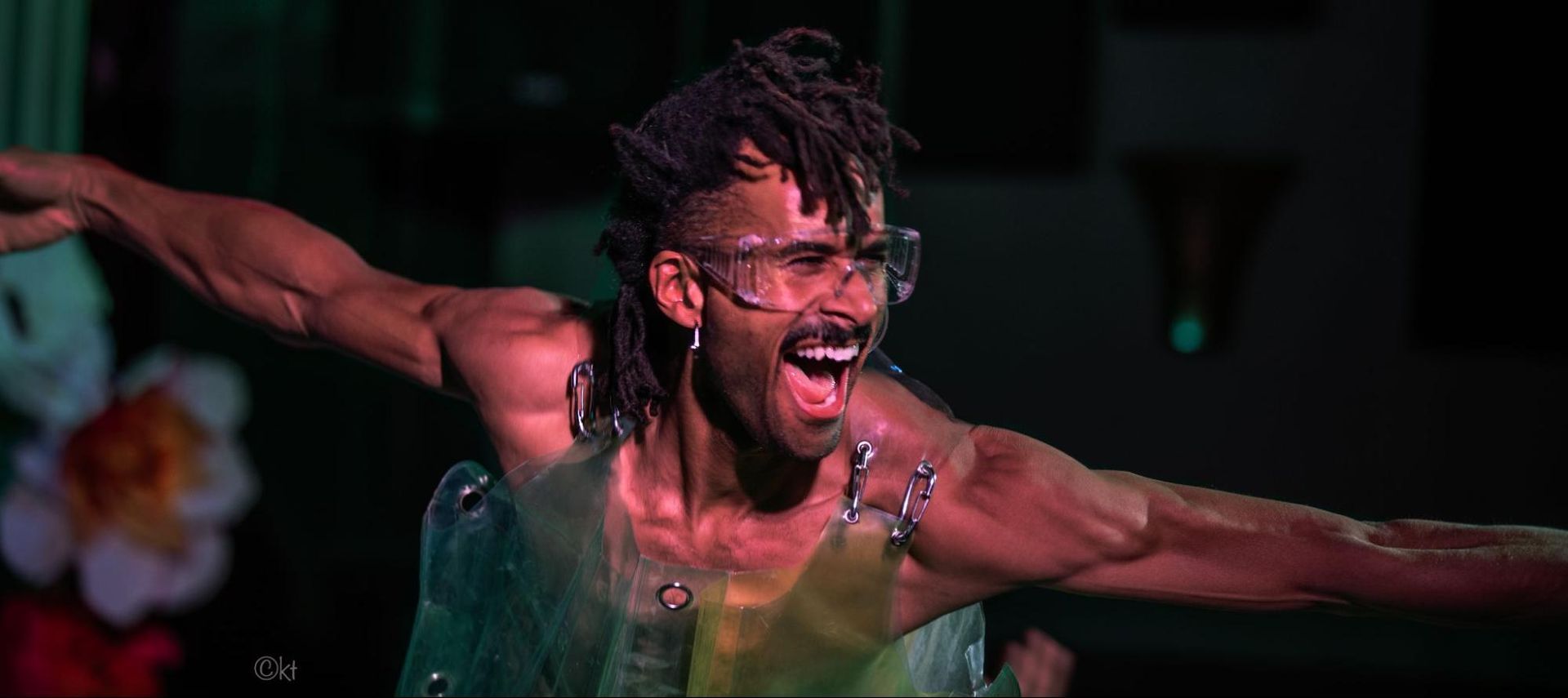 Person in a green costume with outstretched arms, smiling onstage under dramatic lighting.