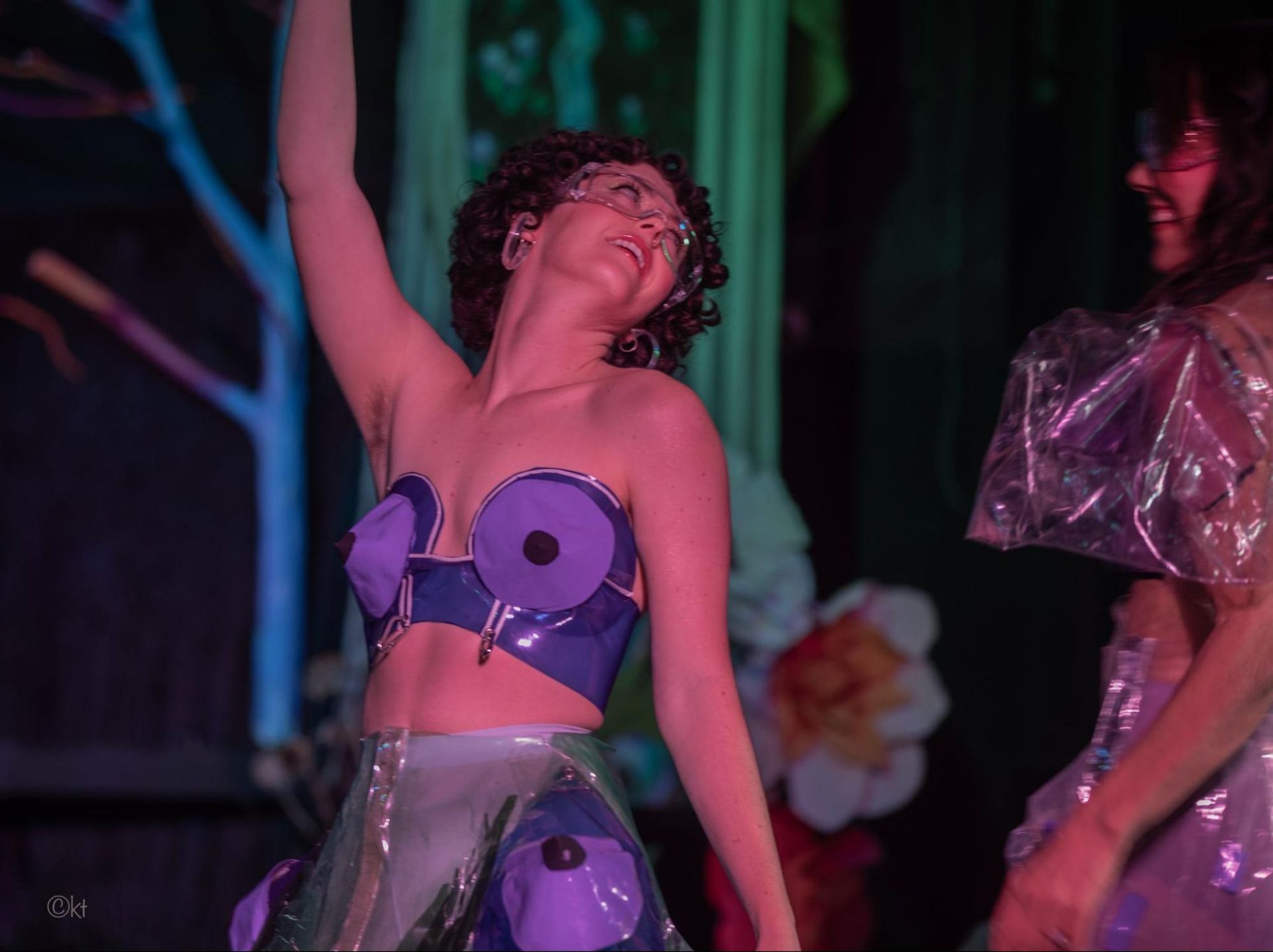 Dancer in a purple costume raises an arm on stage under colorful lighting.