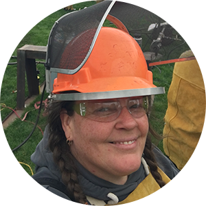 Smiling person wearing an orange hard hat, safety glasses, and yellow protective gear outdoors.