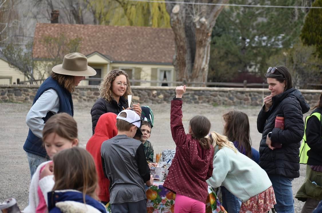 Children gather outdoors while two adults supervise, one holding a camera in a yard with houses and trees.