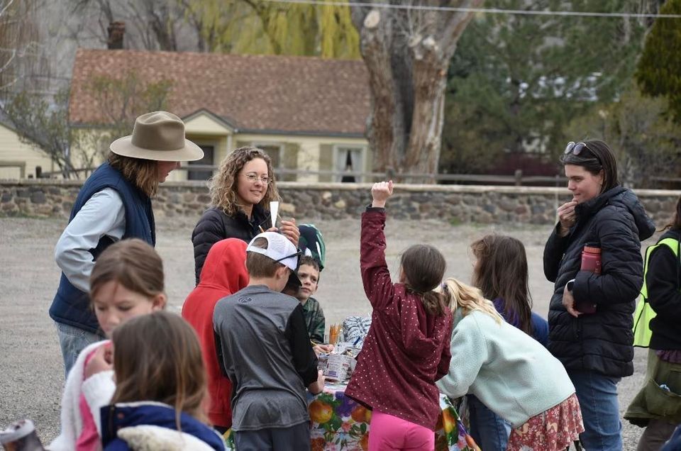 Children gather outdoors while two adults supervise, one holding a camera in a yard with houses and trees.