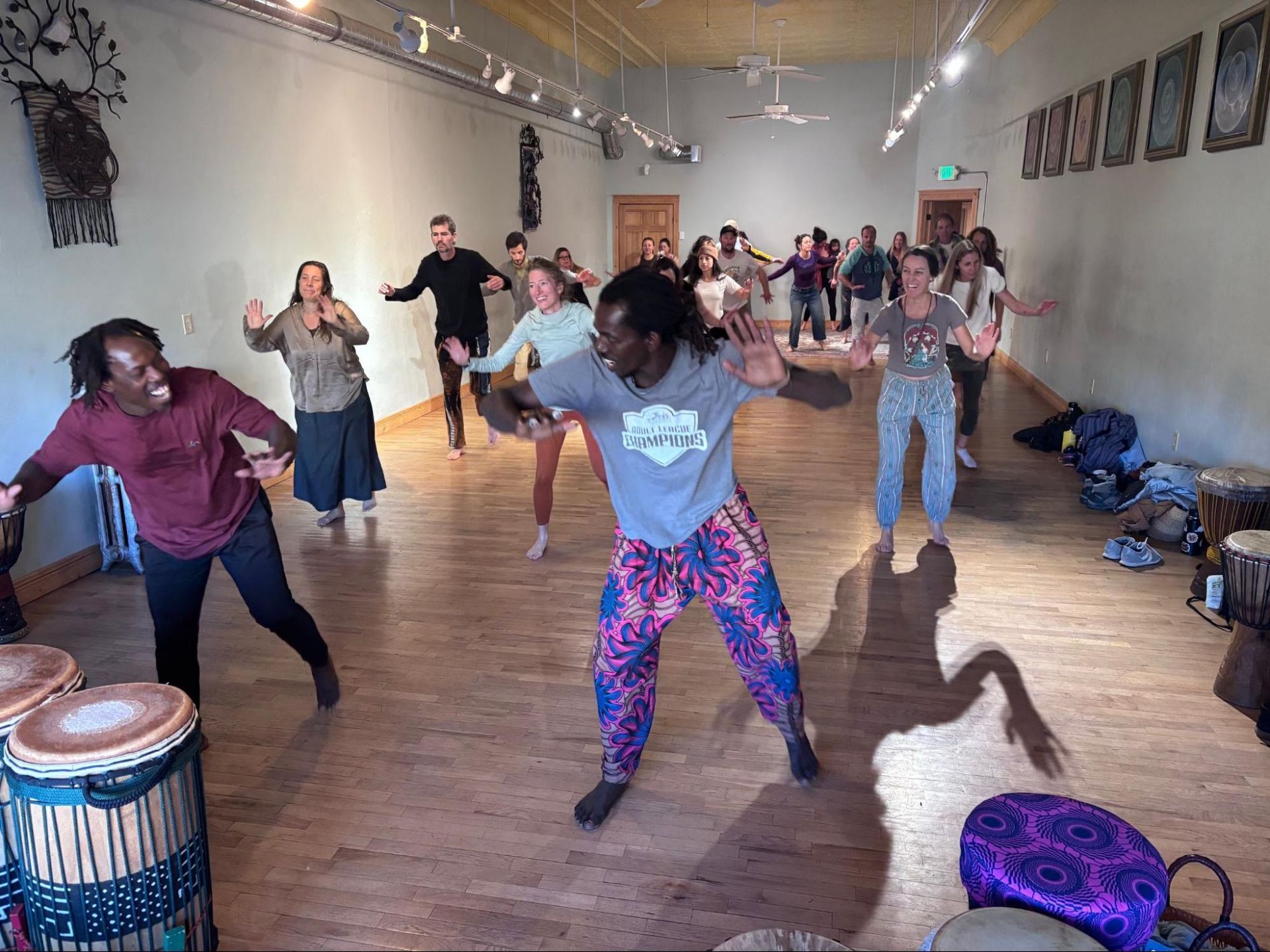 People dancing in a studio with wooden floors, drums, and colorful clothing