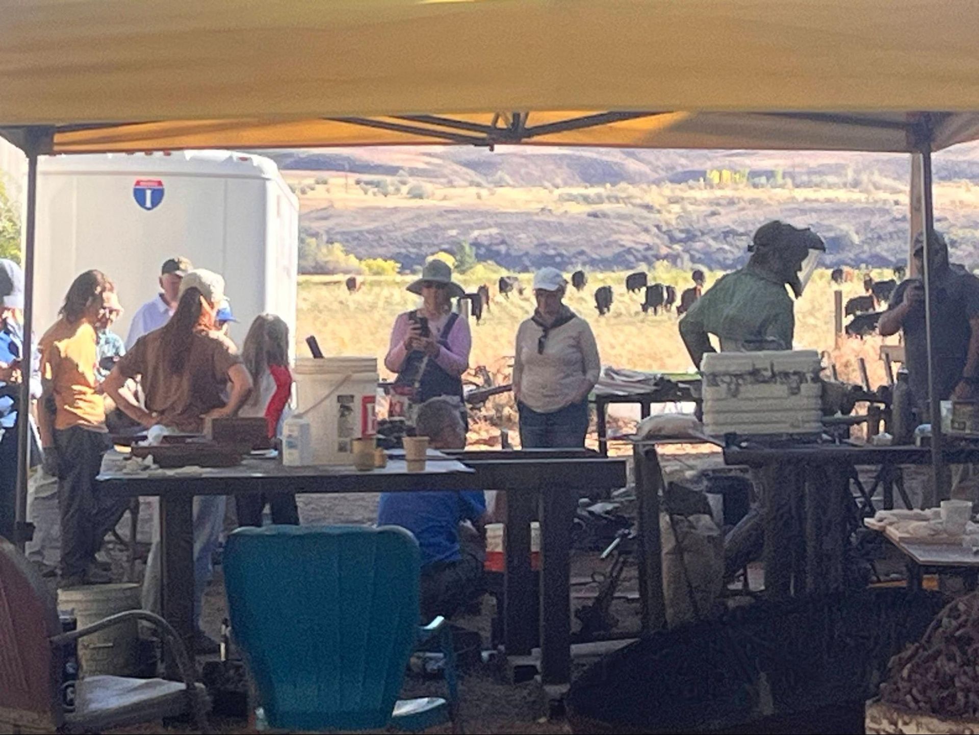 People under a canopy at an outdoor market or worksite, with tables, gear, and hills in the background.